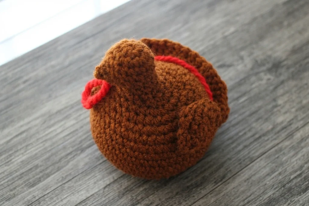 Brown crocheted turkey body with a red wattle and a raised tail section, resting on a grey wood-grain surface, shown without beak, eyes or feathers.