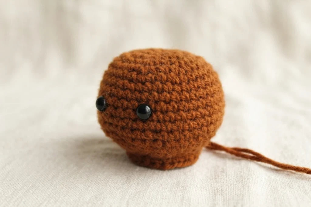 Crocheted dark brown turkey head and upper body with two black safety eyes, resting on a light textured fabric with a loose strand of brown yarn extending to the right.