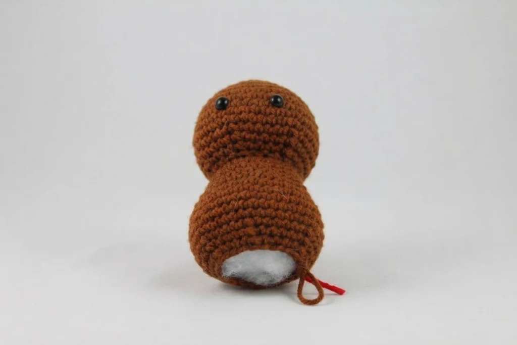 Crocheted brown amigurumi turkey head and body in progress, two black safety eyes, white stuffing showing at the open base, a brown yarn tail and a short red thread on a plain light background.