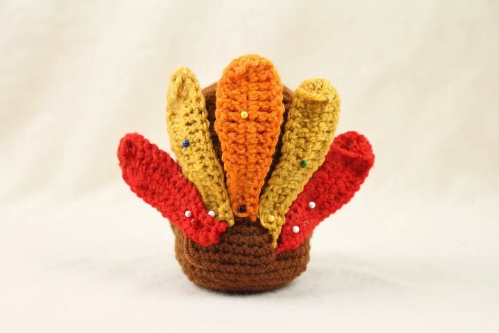 Crocheted brown turkey body with five pinned feathers in orange, gold and red, held in place with colored pins, with loose yarn tails visible against a plain background.