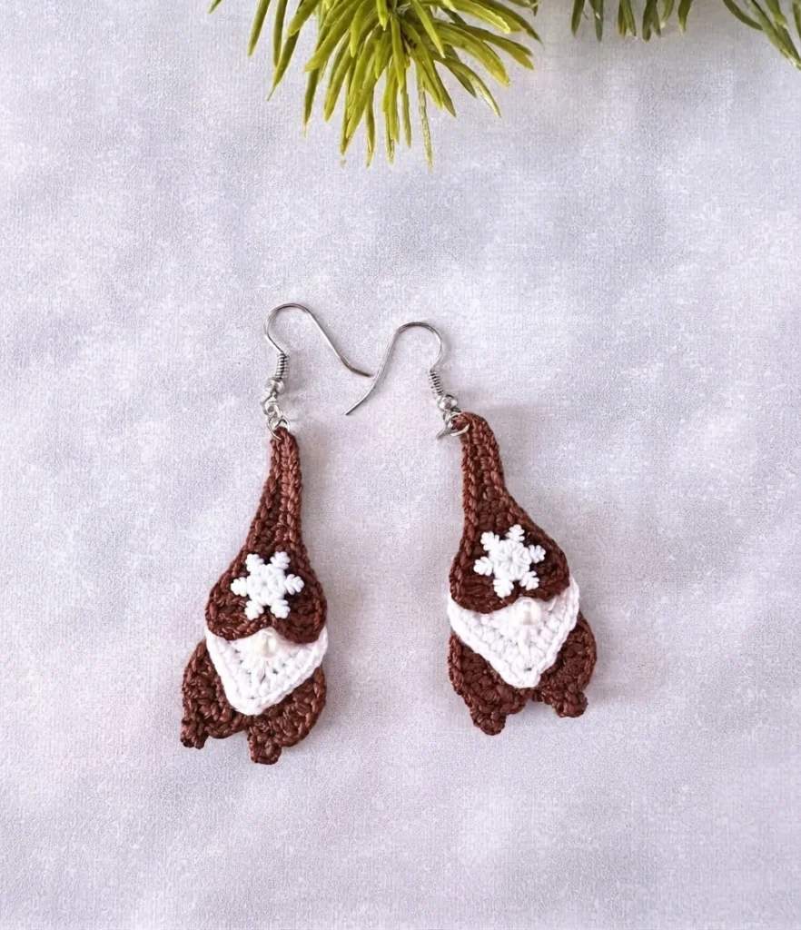 Two crocheted gingerbread gnome earrings in brown with white beards, small white snowflake buttons and pearl noses, hanging from silver ear wires on a light textured surface with green pine sprigs at the top.