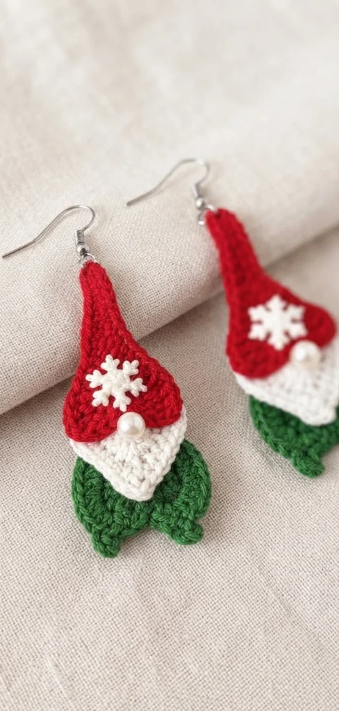 Two crochet gnome earrings with red pointed caps, white snowflake buttons, white heart-shaped beards with pearl noses and green bodies, hanging from silver ear wires on a beige fabric background.