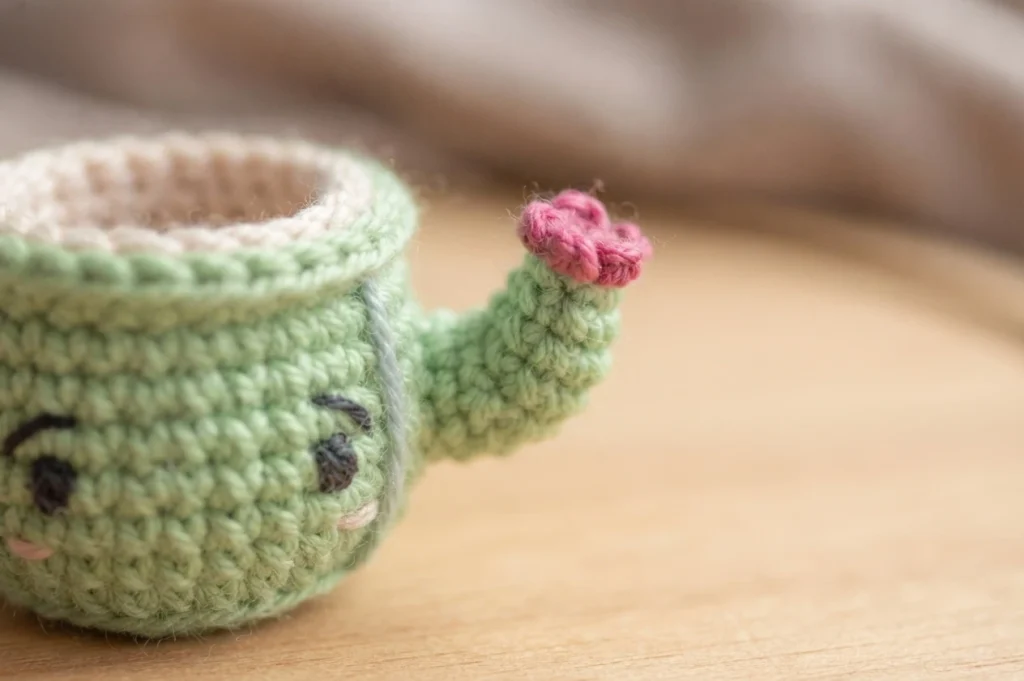 Taza cactus amigurumi verde de crochet con bracito y flor rosa, vista de cerca sobre superficie de madera