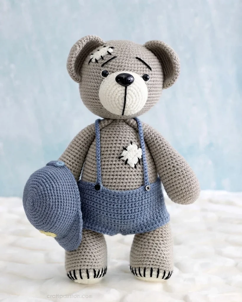 Crocheted grey teddy bear wearing blue suspender pants and holding a blue crocheted cap, with stitched mending patches on its forehead and chest