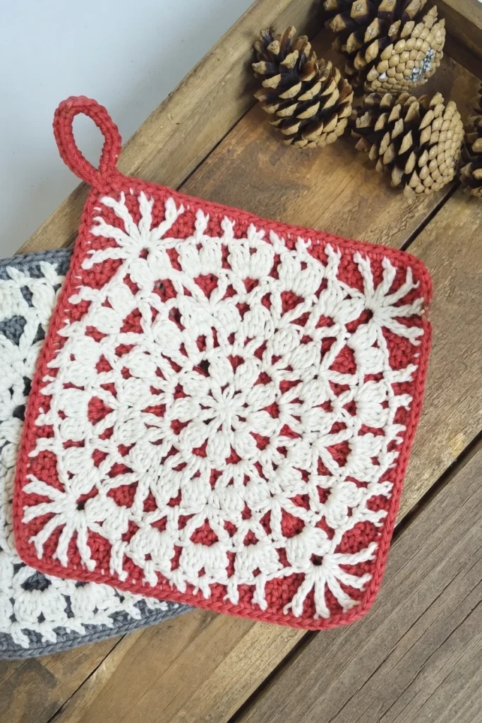 A red and white floral crochet potholder with a hanging loop rests on a wooden surface, partly covering a gray and white potholder.