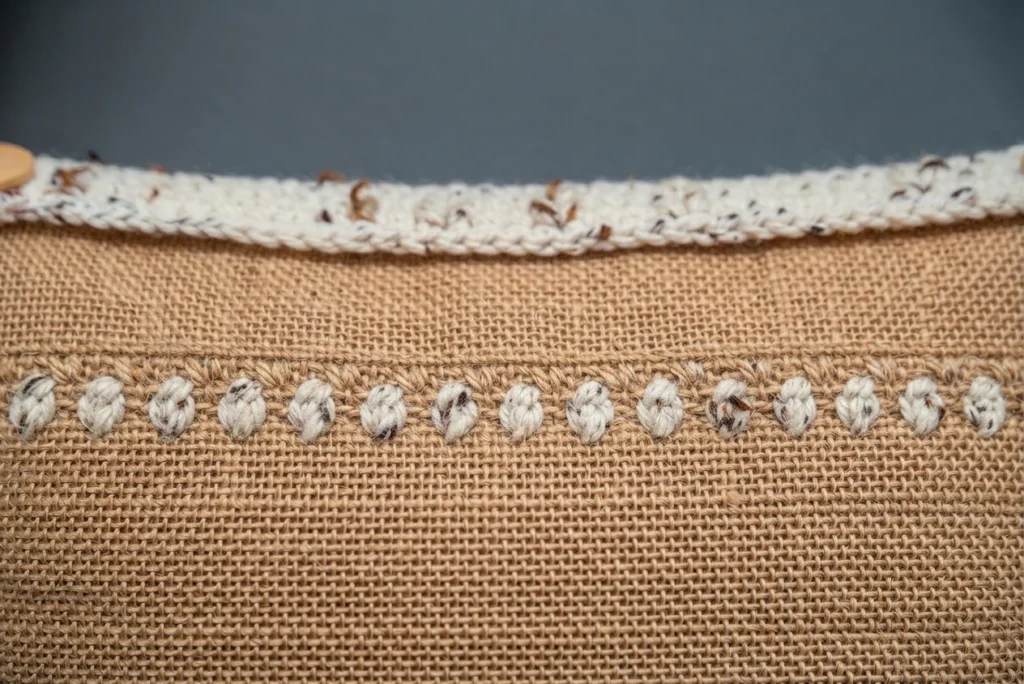 Close-up of a burlap panel with a horizontal row of small crocheted knots and a cream tweed crochet edge running along the top, with a wooden button partly visible at the upper left