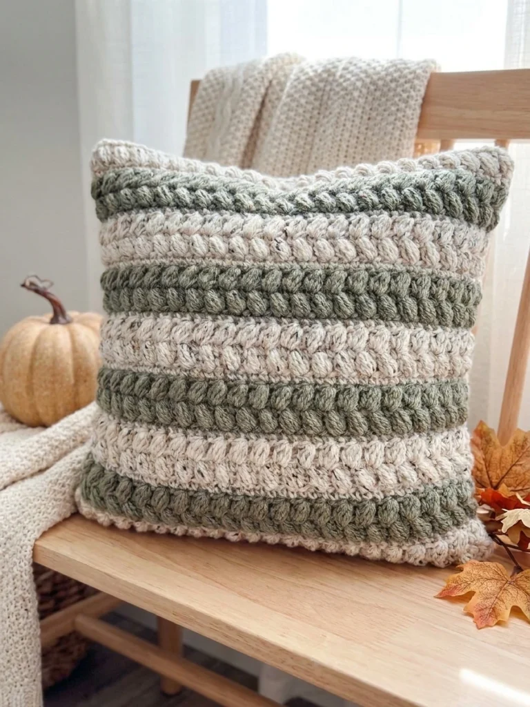 A crocheted pillow with alternating sage green and cream horizontal stripes resting on a light wooden bench, with a small pale pumpkin, dried autumn leaves and a cream knit blanket nearby.