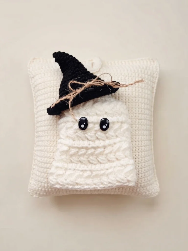 Coussin en crochet crème avec un fantôme blanc appliqué sur le devant, coiffé d'un chapeau de sorcière noir orné d'un lien en ficelle et de deux yeux noirs brillants, posé sur un fond clair et uni.