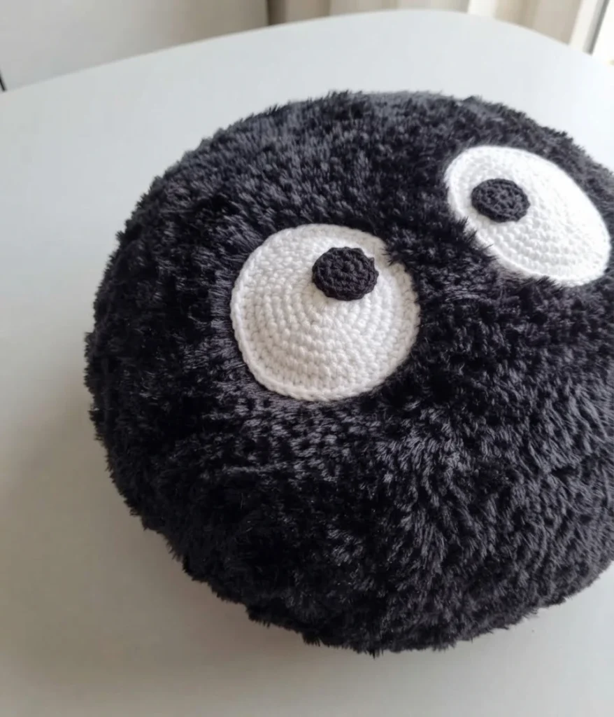 Round black fluffy crochet Soot Sprite pillow with large white eyes and black pupils on a white table