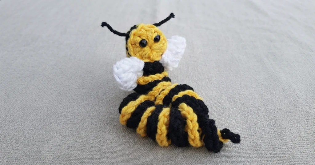 Ver d'inquiétude crocheté en forme d'abeille, corps rayé jaune et noir, ailes blanches, antennes noires et petits yeux brillants, posé sur un tissu gris clair