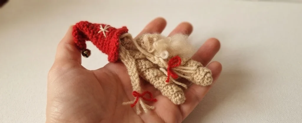 Main tenant un petit worry worm crocheté en forme de gnome de Noël, avec bonnet rouge, clochette, barbe claire et corps beige en spirale