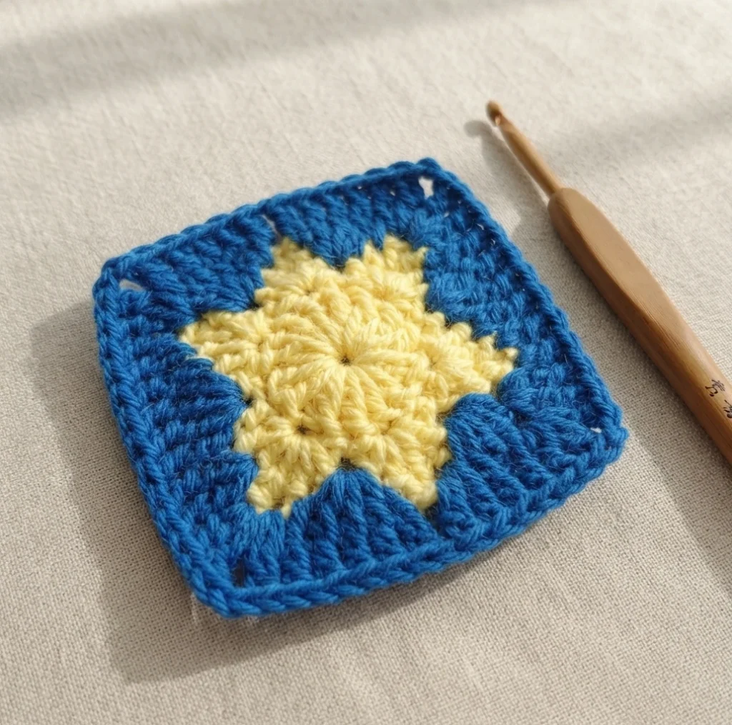 Finished crochet granny square with a pale yellow star center and blue surround, next to a wooden crochet hook on beige fabric