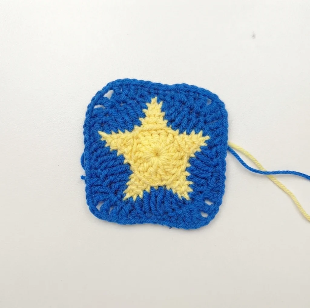 Crochet granny square with a pale yellow star in the center on a royal blue background and loose yarn tails on a white surface