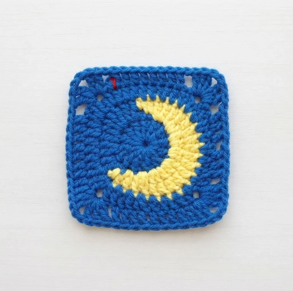 Blue crochet granny square with a yellow crescent moon motif in the center on a white surface