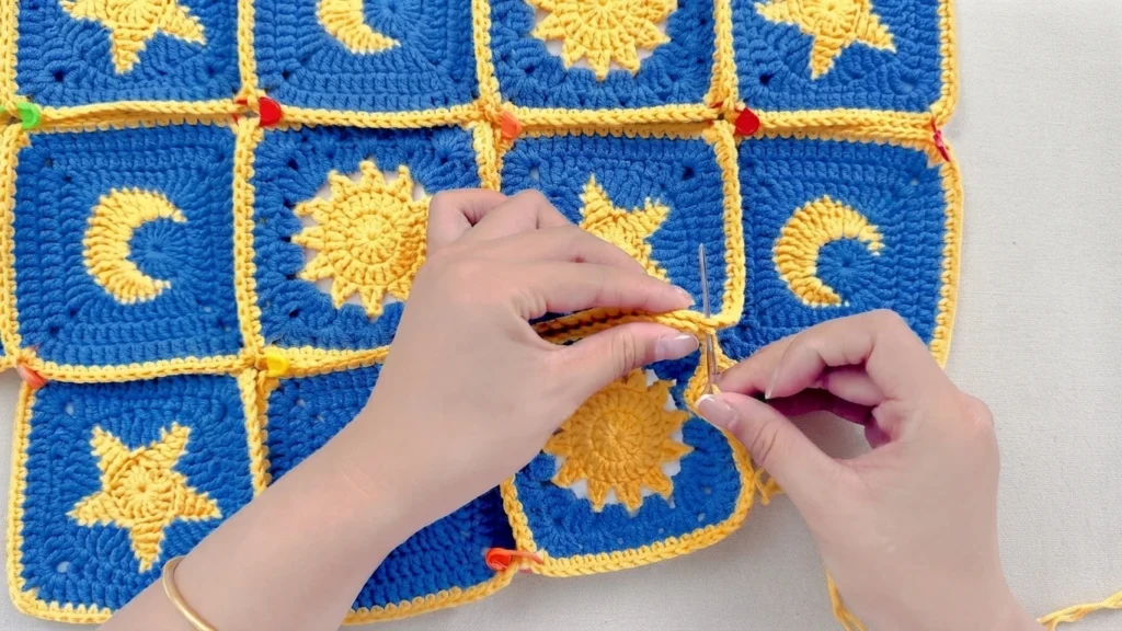 Hands whipstitching blue granny squares with yellow sun, moon and star motifs, held in place with colorful stitch markers