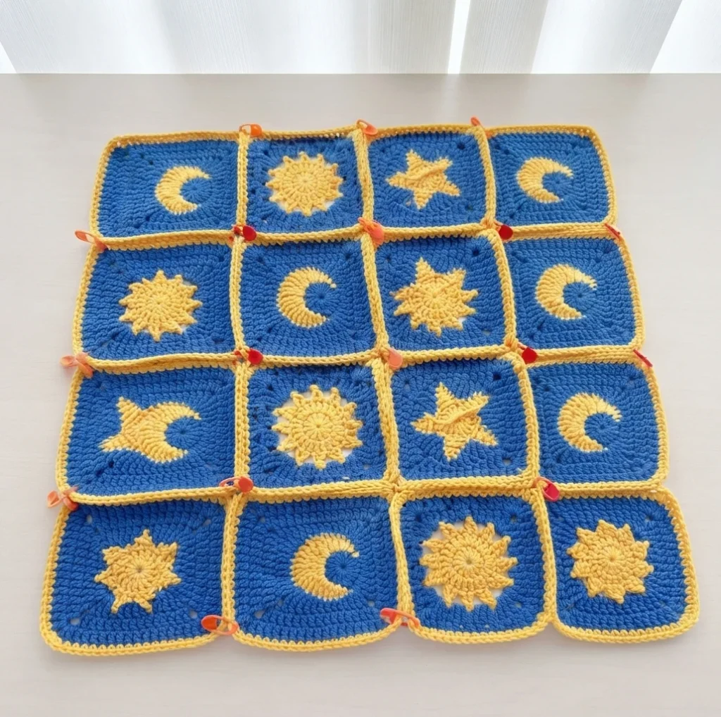 Flat layout of blue granny squares with yellow sun, moon and star motifs, yellow borders and orange stitch markers