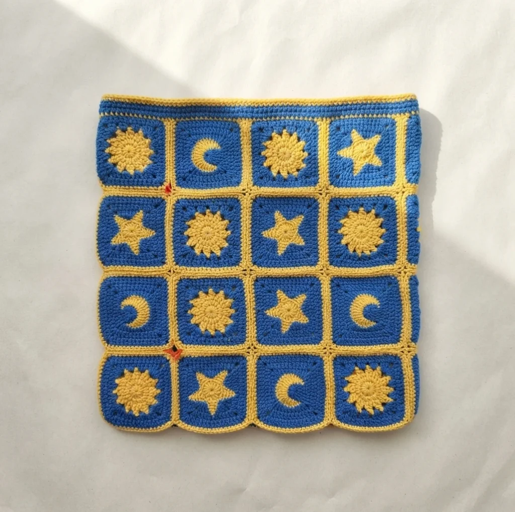Flat panel of royal blue granny squares with yellow sun, moon and star motifs, yellow joining seams and small orange stitch markers