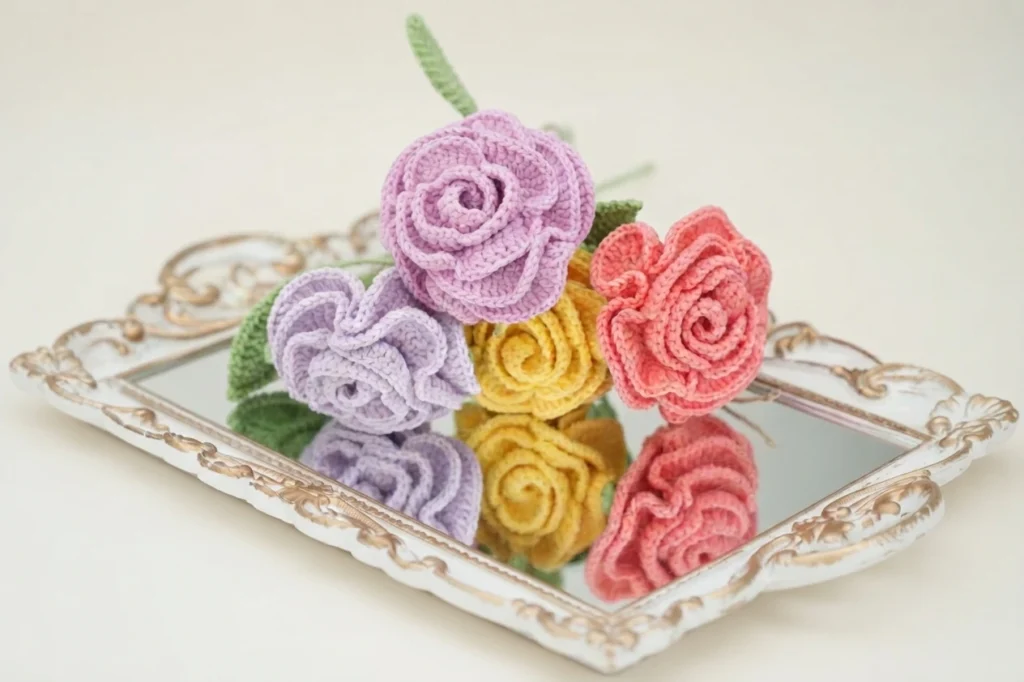 Four crochet roses in pink, lavender, yellow and coral with green leaves on an ornate mirrored tray