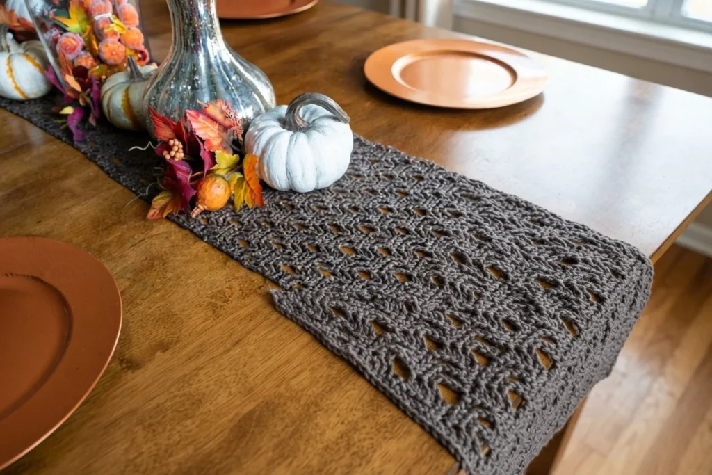 A gray openwork crochet table runner with autumn leaves and pumpkins arranged on a wooden table.