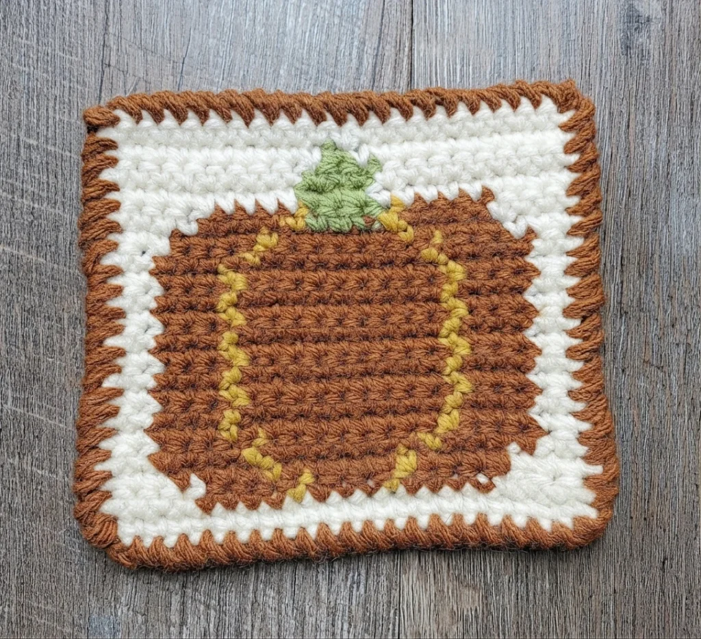 A brown crocheted pumpkin coaster with yellow curved lines, a green stem, a cream background, and a brown border on a wooden surface.