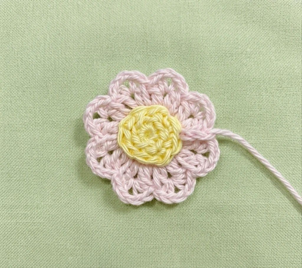 Close-up of a crocheted flower with pink lacy petals around a yellow center, lying on green fabric with a loose thread.