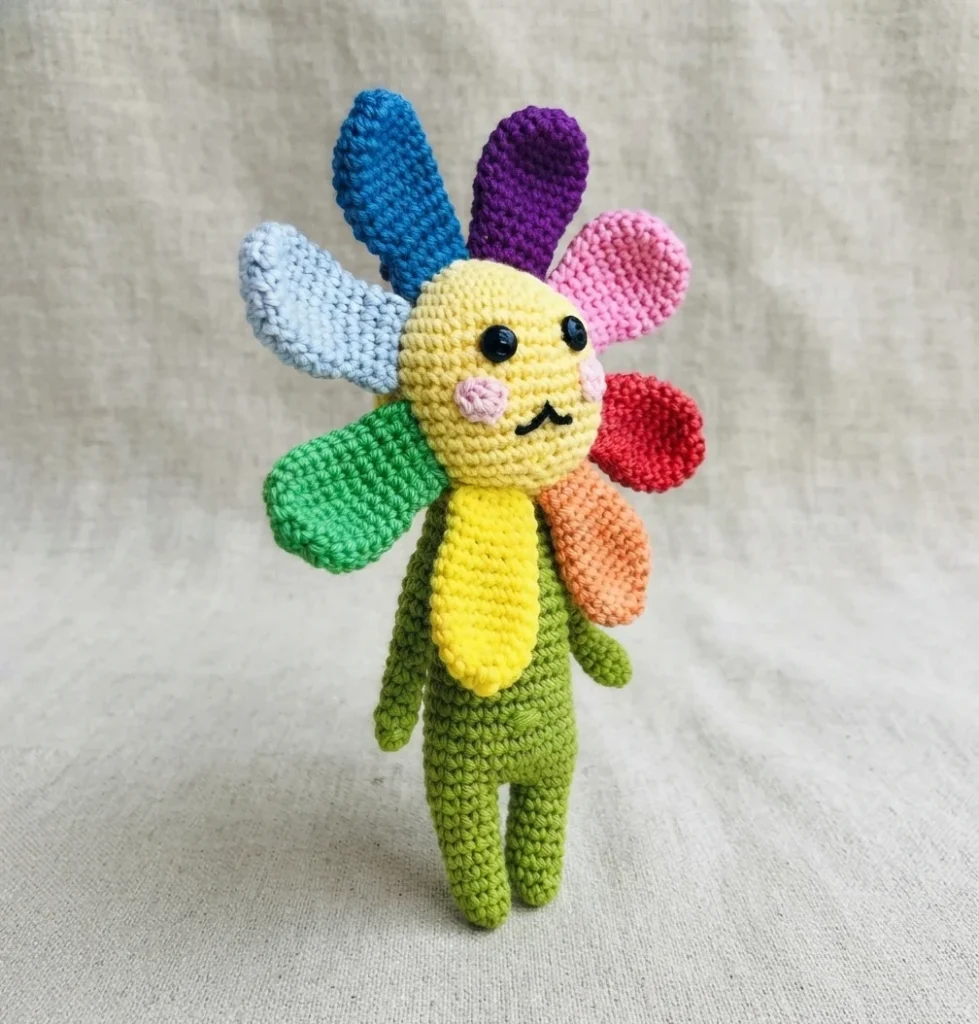 Crochet flower doll with rainbow colored petals, smiling yellow face with rosy cheeks, and green body, standing on a linen fabric background.