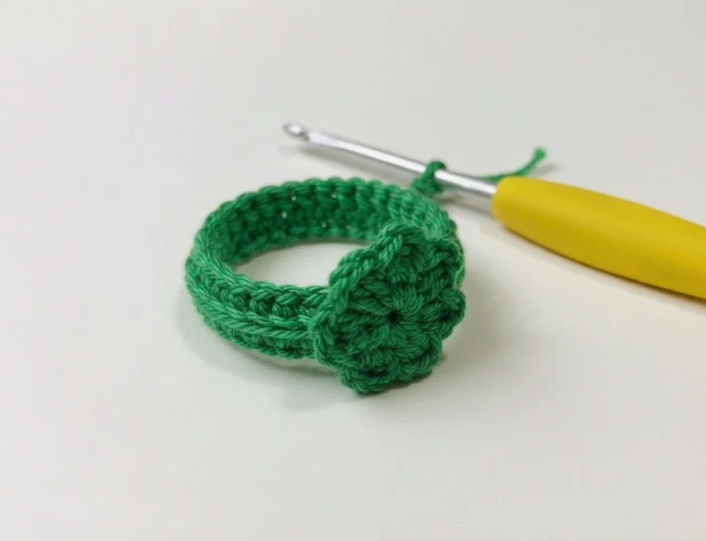 Green crocheted ring with a small green crochet flower, photographed next to a yellow-handled crochet hook on a white background.