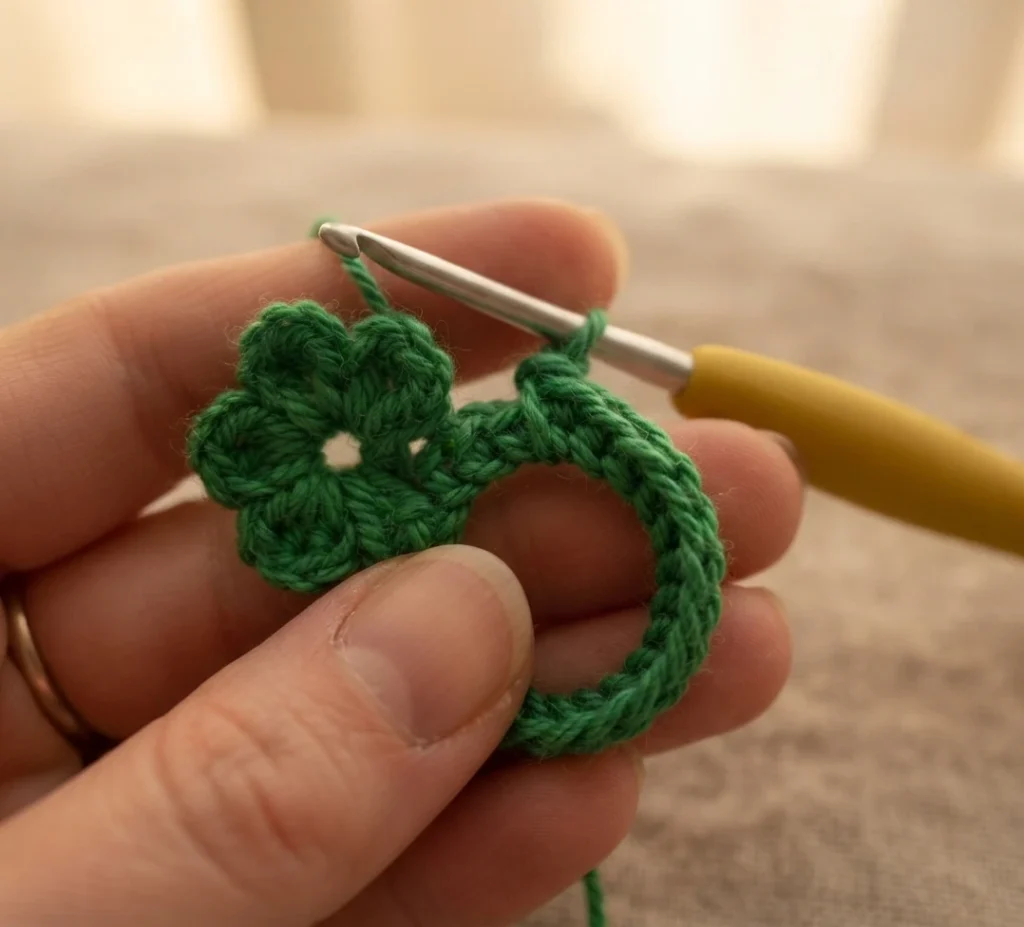 A hand holds a small green crocheted flower ring in progress on a crochet hook.