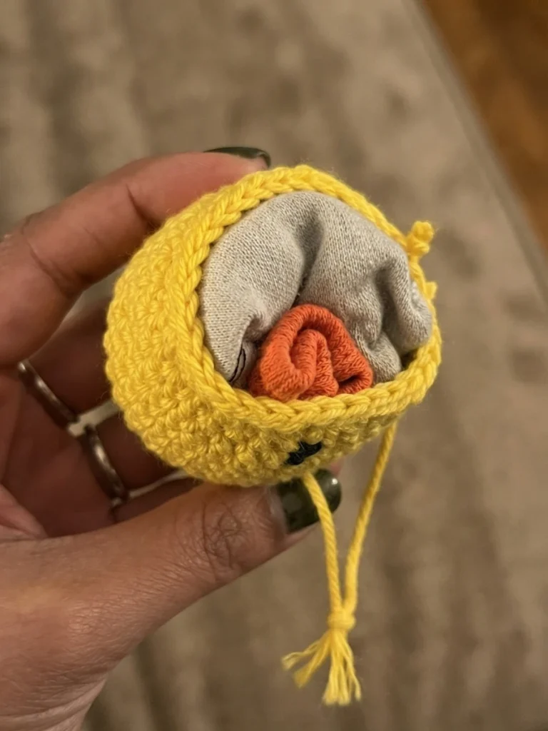 Hand holding an open yellow crochet footbag with a gray sock and orange fabric placed inside as filling, yellow yarn tail hanging down