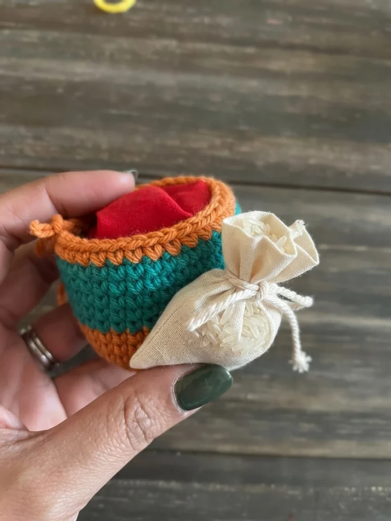 Hand holding an open orange and teal crocheted footbag with a red fabric pouch inside, next to a small white drawstring bag filled with rice.