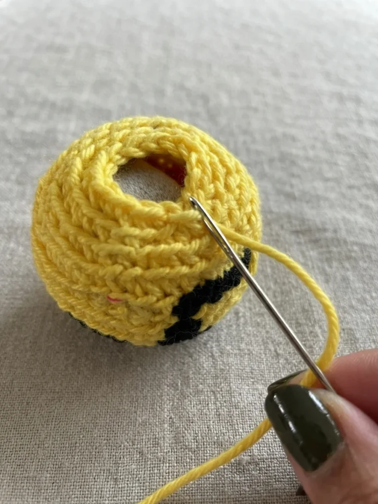 Yellow crocheted footbag ball with its top opening still open, a tapestry needle pulling a long yellow yarn tail through the final rounds of stitches as a hand starts to close it