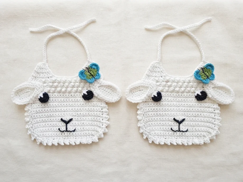 Two finished white crochet lamb baby bibs with floppy ears, black embroidered eyes and mouths, tie straps and blue-green butterfly appliqués, laid flat on cream fabric.