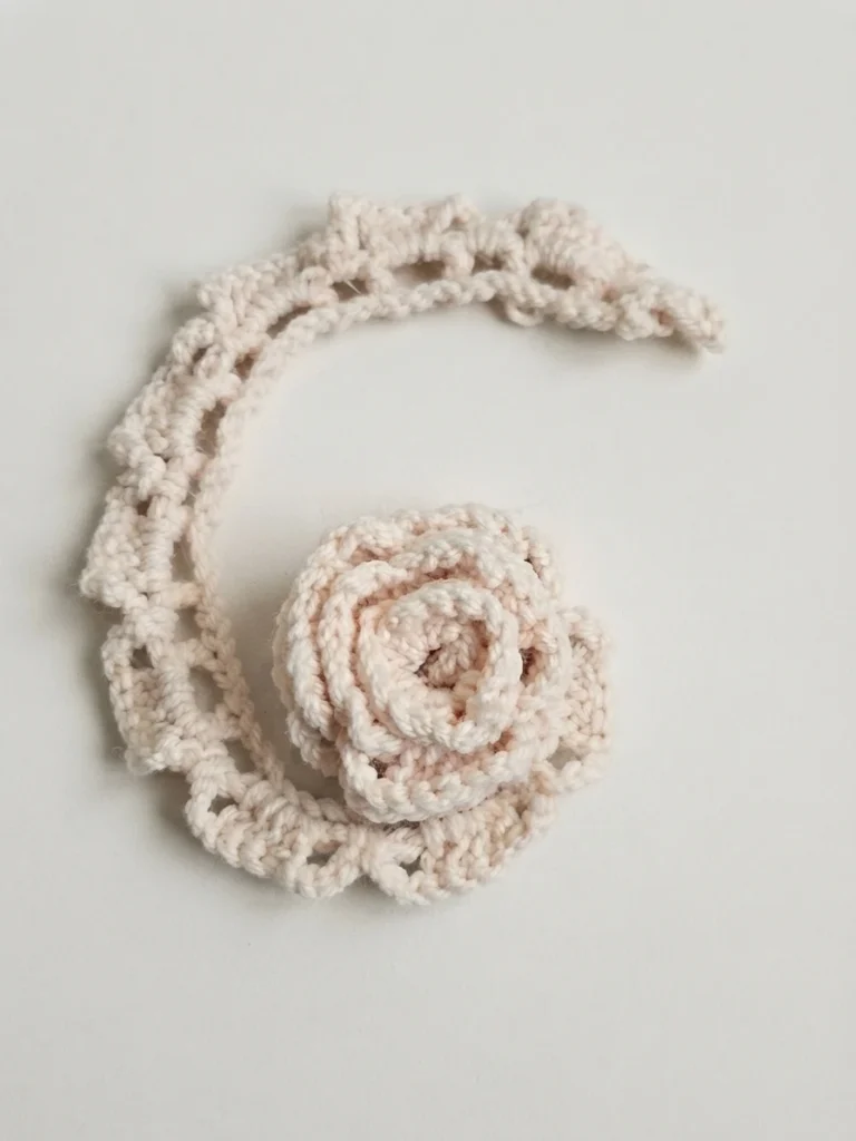 Cream crochet rose beside a partially rolled openwork strip on a plain light background.