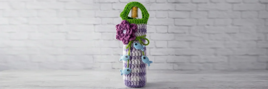 Crochet wine bottle cozy in purple and white plaid with a green handle, decorated with a purple flower and small blue fish appliques on green ties