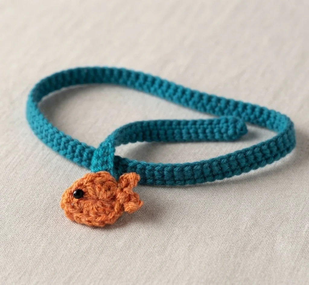Dark blue crochet tie with a small orange crochet fish attached near one end, laid on light fabric.