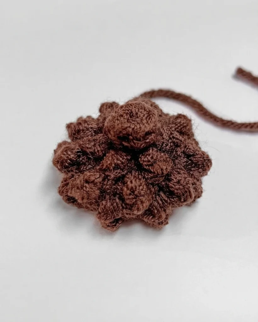 A small brown crocheted cap with raised stitches and a loose yarn tail on a white surface.