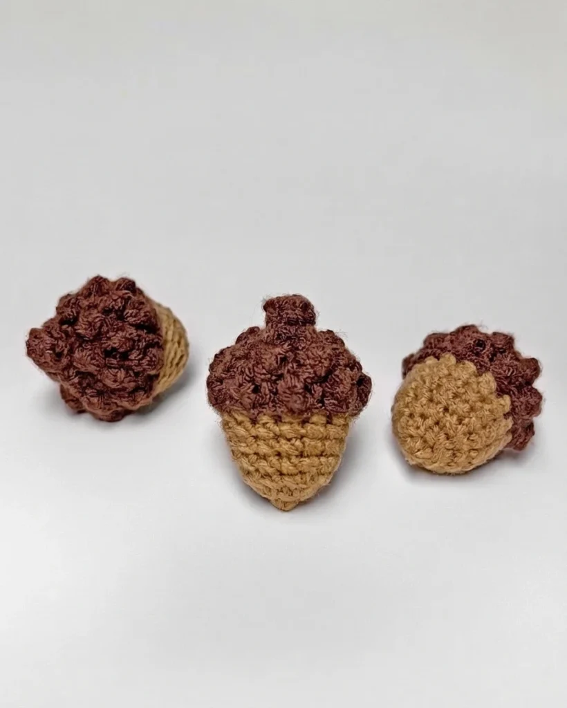 Three small crocheted acorns in tan and brown yarn arranged on a light background.