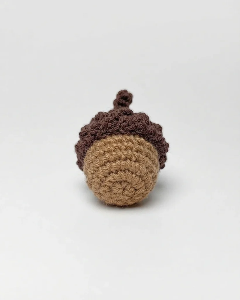 A small tan and brown crocheted acorn with a textured cap and stem on a white background.