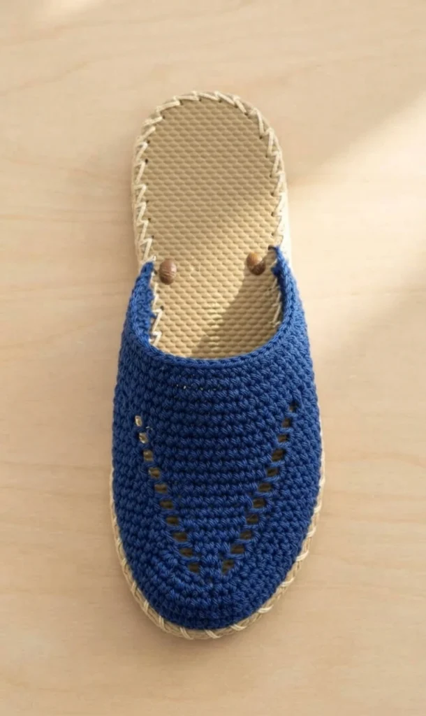 Blue crochet slipper upper with openwork rows stitched to a tan rubber sole with cream whipstitch edging, viewed from above on a wooden surface.