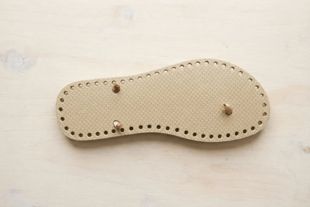 Beige flip-flop sole with small holes punched around its edge, lying on a light wooden surface.