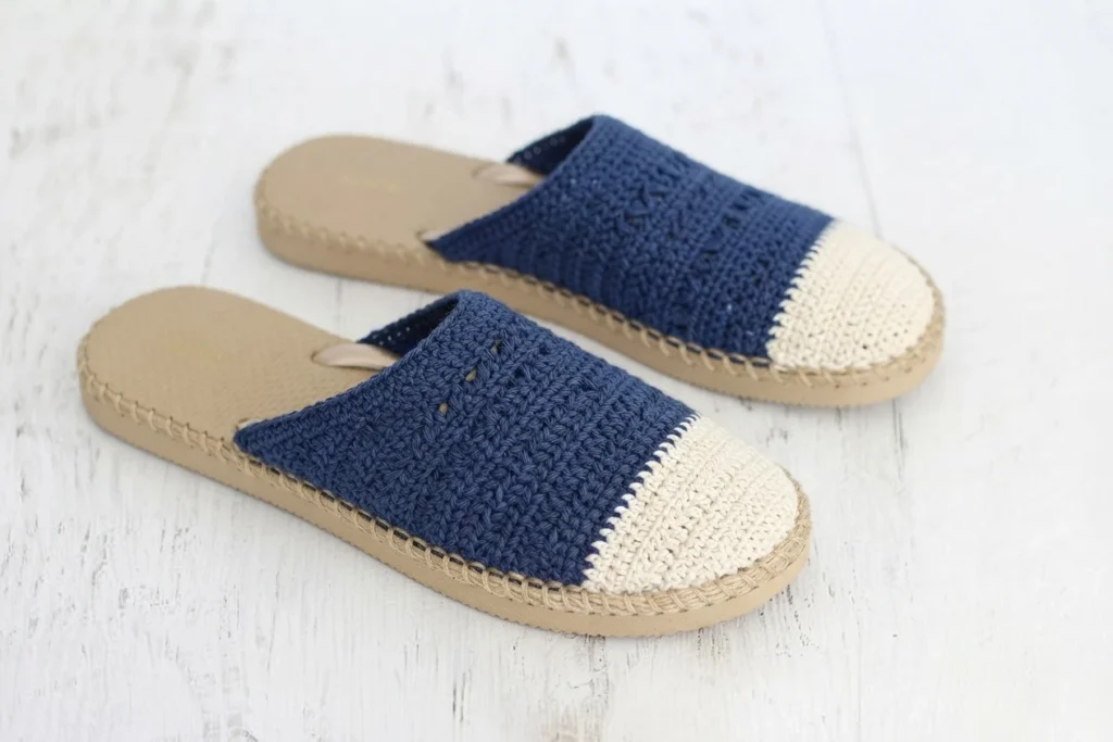 Pair of navy and ecru crochet slip-on shoes with beige rubber soles on a white wooden surface.