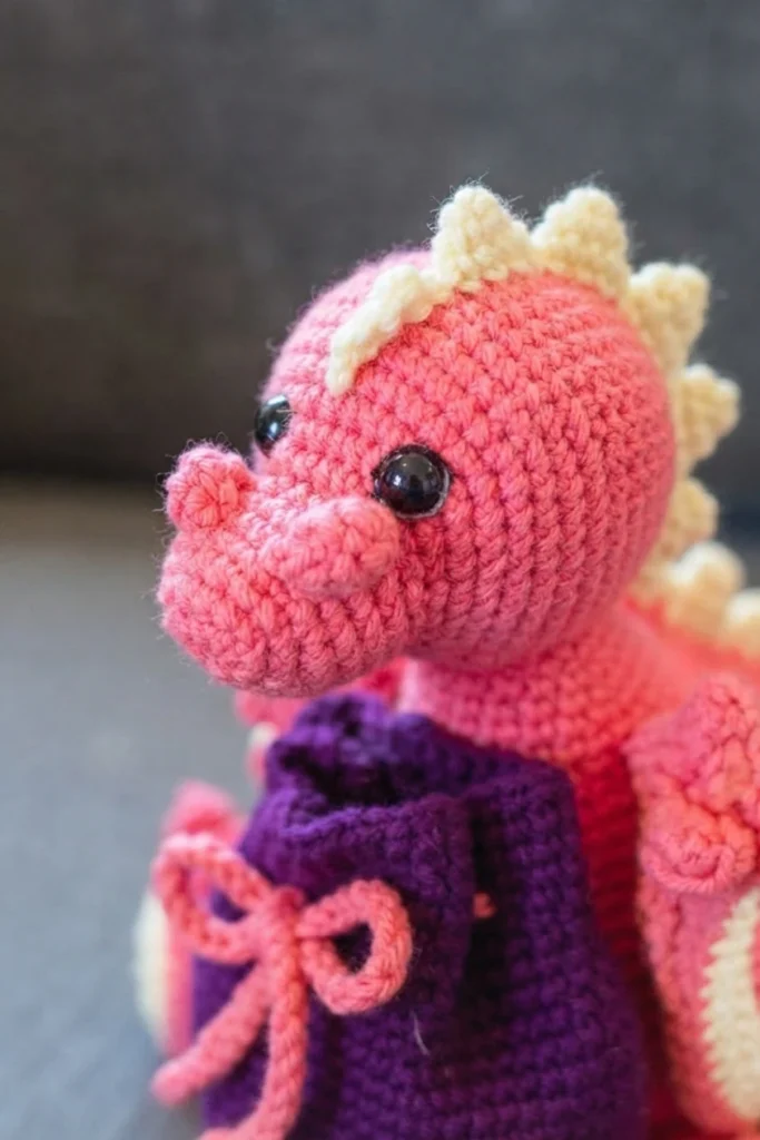 Close-up of a pink crochet dragon with cream spikes and black eyes beside a purple drawstring dice bag with a pink bow