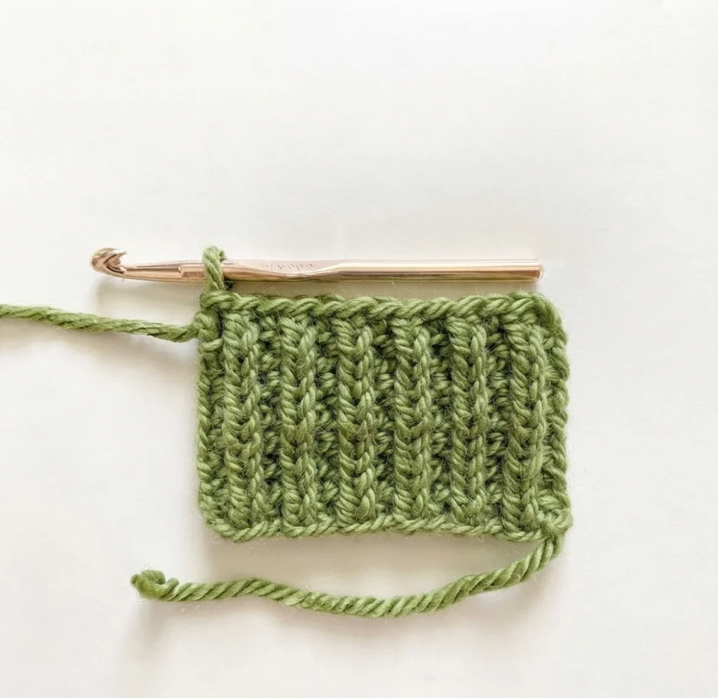 Green crochet swatch with raised vertical posts and a metal crochet hook inserted at the top, on a white background.