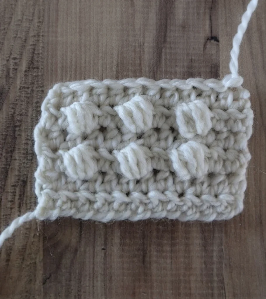 Close-up of a white crochet swatch with six raised bobble stitches arranged in two rows on a wooden surface