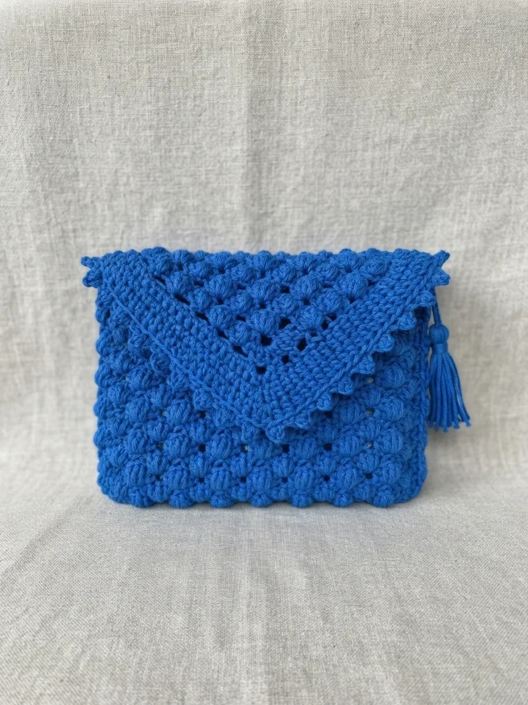 Royal blue crochet clutch bag with textured lotus stitch, pointed flap with picot edging and a side tassel, photographed on light fabric