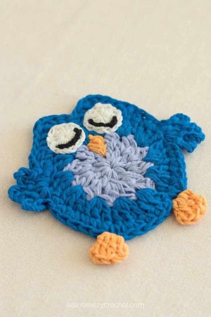 Blue crochet owl coaster with two white eyes outlined in black, an orange beak and orange feet, and a lavender textured center, resting on a plain beige fabric surface with a small site watermark near the bottom.