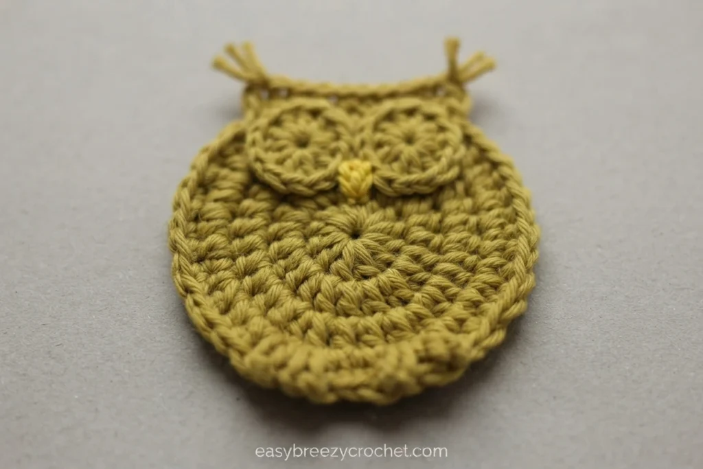 Finished mustard yellow crocheted owl coaster with two round textured eye circles, a small beak and two yarn ends sticking up at the top, photographed on a plain gray background.