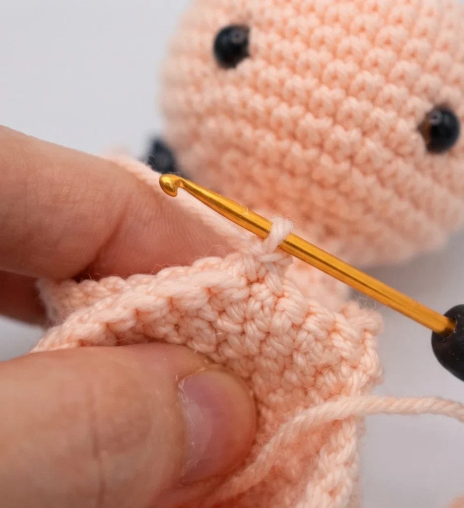 Hand holding a gold crochet hook while working peach yarn in the round on a small crocheted piece, with loose black yarn ends nearby and a peach crocheted ball head with black safety eyes in the background.