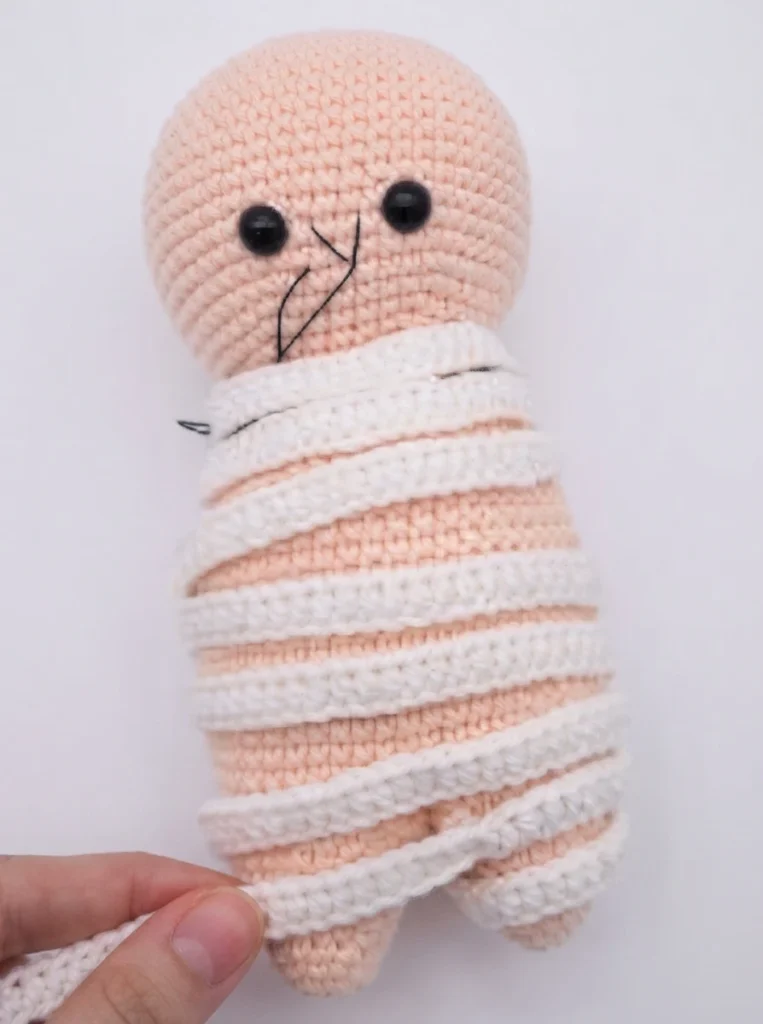 Crocheted mummy amigurumi doll with a peach head, black safety eyes, embroidered black brows and mouth, and white crocheted strips wound diagonally around its body, held by a hand at the lower left against a plain pale background.