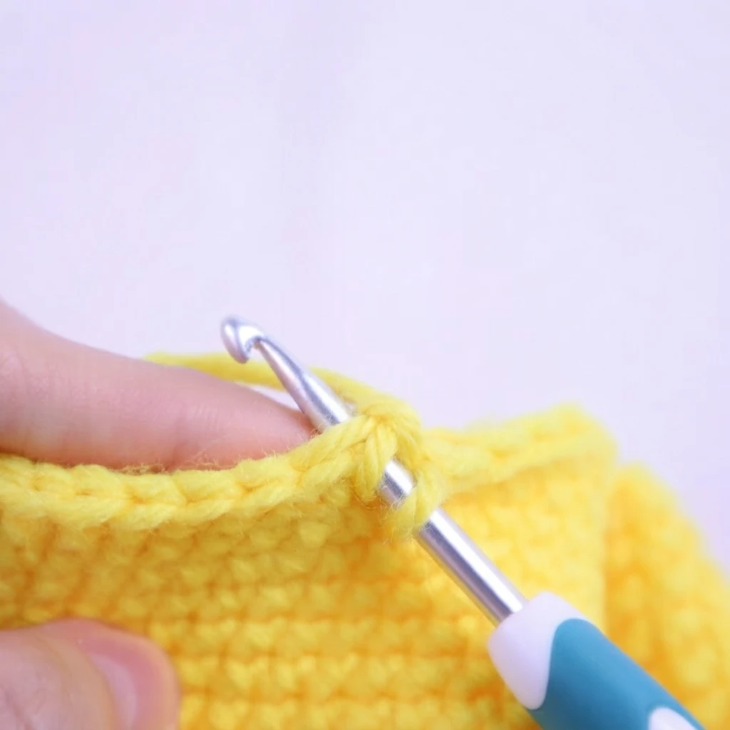 Close-up of a hand holding a yellow crocheted swatch while a teal-handled crochet hook works a stitch, with a loop of yarn on the hook, against a pale lilac background.
